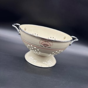 May include: Cream-colored colander with a seashell design and silver trim. The colander has a pedestal base and two silver handles. The design includes a pink seashell and a thin pink stripe. The colander is made of metal.