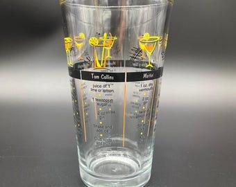 May include: A clear glass cocktail shaker with a black band featuring the text "Tom Collins" and "Martini." The glass has yellow and black illustrations of cocktails and includes drink recipes. The shaker is designed for mixing drinks.