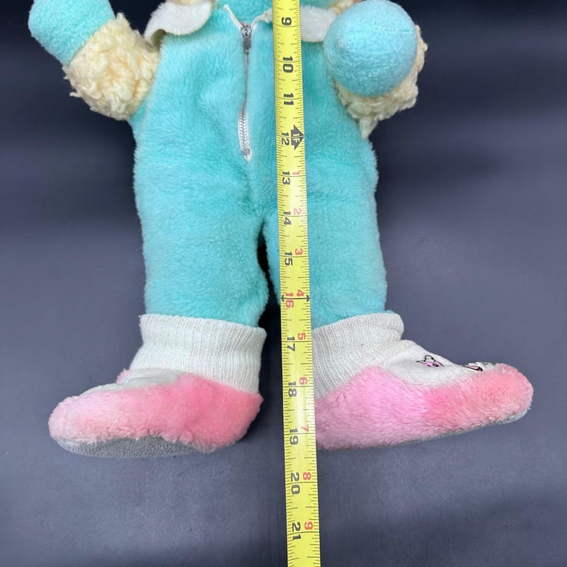 Peut inclure: Une poup&eacute;e en peluche vintage dans une combinaison bleu clair avec une fermeture &eacute;clair, une garniture cr&egrave;me et des chaussons roses. La poup&eacute;e mesure environ 53 cm de haut. Les chaussons ont un revers blanc en forme de chaussette.