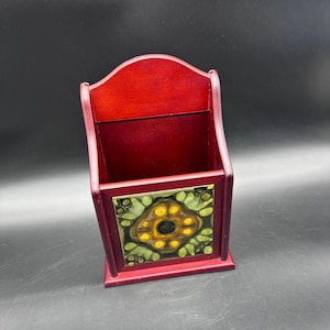May include: A red wooden wall-mounted organizer with a decorative tile on the front. The tile features a floral design with yellow and green accents. The organizer has a curved top and a rectangular opening.