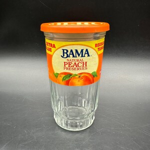 May include: A clear glass jar with an orange lid and a label featuring the "BAMA Natural Peach Preserves" logo. The label has a yellow background with orange and red accents, and images of peaches.