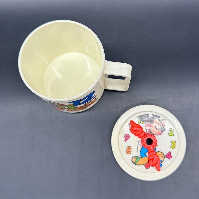 Puede incluir: Taza de pl&aacute;stico color crema con asa y tapa a juego. La taza presenta gr&aacute;ficos de dibujos animados y la tapa tiene un dise&ntilde;o de esfera de reloj con un personaje de dibujos animados y manecillas rojas. La taza est&aacute; vac&iacute;a.