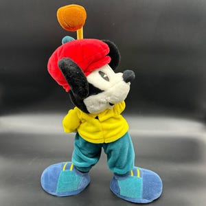 Puede incluir: Un juguete de peluche de Mickey Mouse con una boina roja, una camisa amarilla y pantalones azules. Est&aacute; sosteniendo un palo de golf con una bola marr&oacute;n en la parte superior.