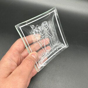 May include: A clear glass trinket dish with a white etched design of two children holding hands. The text "Share with the One you Love" is etched on the bottom of the dish.