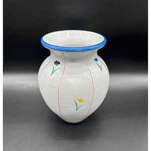 May include: White ceramic vase with a blue rim and floral accents. The vase features vertical pink stripes and small painted flowers in purple, yellow, and blue. A decorative piece for home decor.