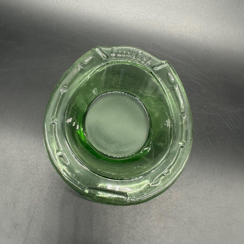 May include: A green glass ashtray with a circular center and a rounded, oval shape. The ashtray has a textured rim and the text "Good Luck" is visible on the rim.