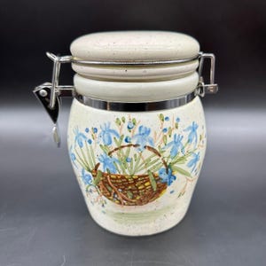 May include: A ceramic jar with a lid and metal clasp. The jar is off-white with a hand-painted design of a basket of blue flowers. The lid and base have a speckled finish. The jar is designed for storage.