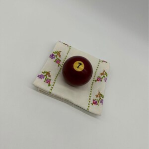 May include: A red billiard ball with the number 7 on it, resting on a white cloth with a floral pattern.