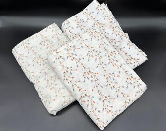 Puede incluir: Cuatro cuadrados de tela blanca doblados con un delicado estampado floral. El patrón presenta tallos marrones con pequeñas flores azules, naranjas y moradas. La tela está dispuesta sobre una superficie oscura.