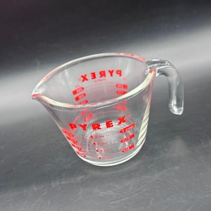 May include: A clear glass measuring cup with a handle and a pouring spout. The exterior features red printed markings including the brand name PYREX, with measurements in cups, ounces, and milliliters for precise liquid volume tracking.