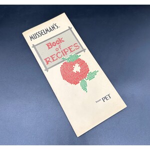 May include: A vintage cookbook titled "Musselman's Book of Recipes" with a cross-stitch style tomato illustration. The cover is cream-colored with the text in black and red. The cookbook is from PET.