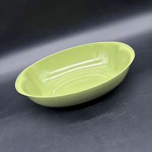 May include: A light green, oval-shaped serving bowl. The bowl has a smooth, glossy finish and gently sloping sides. The interior is a uniform light green color, and the bowl appears to be made of a durable material. The bowl is empty and sits on a dark surface.
