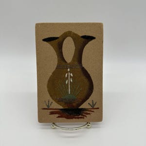 Puede incluir: Una pintura de arena marrón que representa un jarrón de cerámica tradicional nativo americano con detalles en blanco y verde. El jarrón está sobre un fondo marrón con hierba verde.