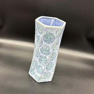 Könnte beinhalten: Eine hellblaue Keramikvase mit sechseckiger Form und einer strukturierten Oberfläche. Die Vase hat ein Muster aus sich überlappenden Kreisen.