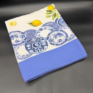 May include: A folded fabric tablecloth featuring a cream-colored background with a pattern of yellow lemons, green leaves, and various blue and white decorative plates with intricate floral and classical portrait designs, finished with a solid blue border.
