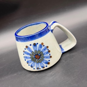 Peut inclure: Une tasse en céramique avec une base de couleur crème, ornée d'un motif floral bleu peint à la main sur le côté. La tasse présente un bord bleu épais et une large anse angulaire. Le motif floral se compose de pétales bleus autour d'un centre.