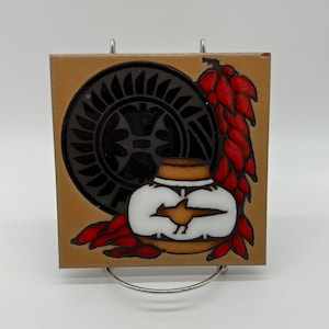 May include: A ceramic tile with a brown background features a black and white stylized design of a pot with red chili peppers. The pot has a white body with a black bird design and a brown rim.