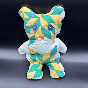 May include: A handmade stuffed animal bear with a patchwork design. The bear features a geometric pattern in green, yellow, and white fabric. It has blue button eyes and an orange button nose. The bear's body is white, and it has a red accent on one paw.