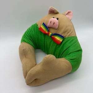 Könnte beinhalten: Ein kissenförmiges Schwein mit einem beigefarbenen Körper, einem grünen Hemd und beigefarbenen Beinen. Das Schwein hat eine rosa Schnauze und Ohren sowie eine Regenbogen-Schleife. Das Kissen ist als Dekorationsartikel konzipiert.