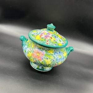 Puede incluir: Un tazón de servir de cerámica verde con tapa, decorado con un patrón floral en tonos de amarillo, rosa, azul y verde.