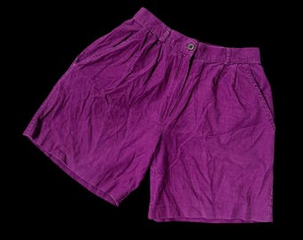 May include: A pair of purple shorts with a button closure and pockets. The shorts are made of a textured fabric and have a high waist. The shorts are displayed against a black background.