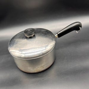 May include: A stainless steel saucepan with a polished lid and a black handle. The lid features a small, round black knob in the center. The handle is made of black plastic and includes a small metal loop at the end for hanging storage.