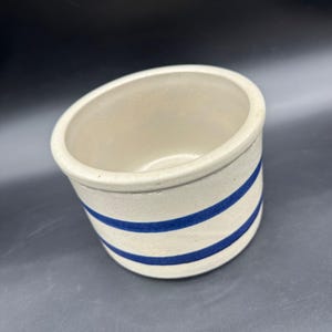 Puede incluir: Un tarro de cerámica de color crema con dos rayas azules horizontales. El recipiente de tapa abierta tiene forma redondeada y un borde grueso. El tarro probablemente se utiliza para almacenamiento o decoración.