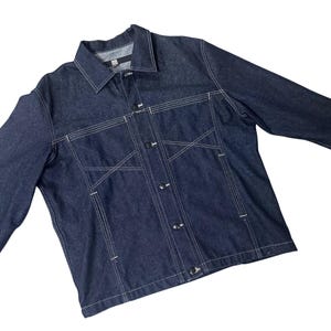 May include: A dark blue denim jacket featuring a button-up front, a collared neckline, and white stitching. The jacket has two chest pockets and long sleeves. The white stitching forms a geometric pattern.