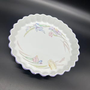 以下が含まれることがあります： ピンク、黄色、紫の花が描かれた、波打つ縁取りのある白いセラミック製のベーキングディッシュ。ディッシュは薄い紫色の縁取りが施されています。