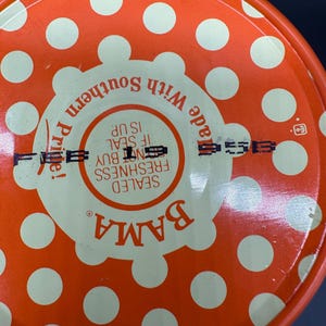 May include: Close-up of a round lid with an orange polka dot pattern. The lid features the word "BAMA" in orange, and text that reads "SEALED FRESHNESS, IF SEAL IS UP, DO NOT BUY". The lid also has the words "Made With Southern Pride!"