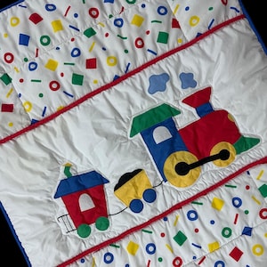 May include: White baby blanket with a colorful train applique. The blanket features a repeating pattern of red, yellow, blue, and green shapes and circles. The train is red, blue, yellow, and green with black wheels and a black smokestack.