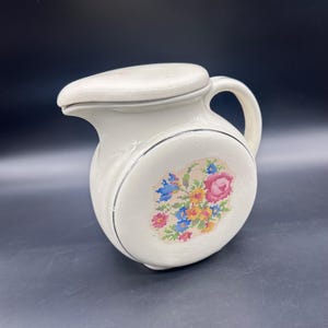 May include: Cream-colored ceramic pitcher with a lid and handle. The pitcher features a cross-stitch floral design in pink, blue, yellow, and green. A thin black line encircles the floral design. The pitcher is on a dark background.