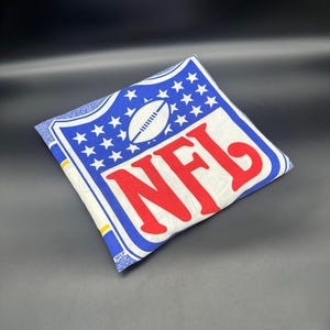May include: A blue and white fabric pillow with a red NFL logo and a white football in the center. The logo is surrounded by white stars on a blue background.