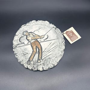 May include: A round, white, textured ceramic wall plaque with a golfer swinging a club. The plaque has a brown and white image of the golfer and a tag that says "Signs of Clay".