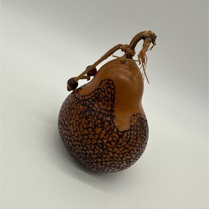 May include: A handcrafted gourd-shaped container with a natural brown finish. The gourd features a detailed, dark brown, scale-like pattern on the lower half. A leather cord with wooden beads and a dried stem complete the design.