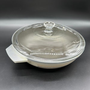 May include: A white ceramic casserole dish with a glass lid. The dish has a black and gold stripe design around the rim.