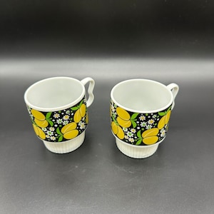 Puede incluir: Dos tazas de cerámica blanca con diseños de frutas y flores negras y amarillas. Las tazas tienen una banda blanca alrededor de la parte inferior.