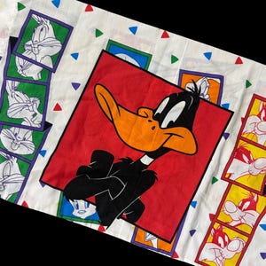 May include: White fabric featuring cartoon characters. A large red square showcases Daffy Duck, while smaller squares display Bugs Bunny and other Looney Tunes characters. The fabric is decorated with colorful triangles.