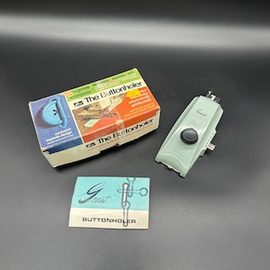 Puede incluir: Un ojalador vintage de plástico azul claro con una perilla negra y la palabra "Greist" impresa en él. El ojalador está empaquetado en una caja con el texto "The Buttonholer" y "accesorio de puntada de ojal automático".