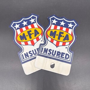 May include: Two shield-shaped metal signs featuring a blue top section with three white stars, a red and white striped middle, and a yellow oval containing the text M-F-A in black. The bottom white section displays the word INSURED in blue letters.