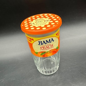 May include: A clear glass jar of BAMA Natural Peach Preserves with an orange lid and label. The label features the brand name, peach illustrations, and the words "Extra Value". The lid has white polka dots.