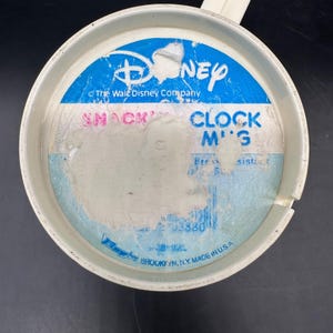 Puede incluir: Una taza de reloj de aperitivos de Disney de pl&aacute;stico blanco con asa. La taza presenta un logotipo de Disney azul y blanco y el texto "SNACK CLOCK M'G". La taza est&aacute; hecha en los EE. UU.