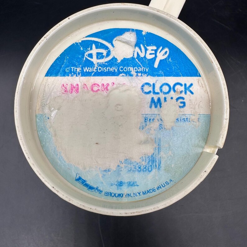 Puede incluir: Una taza de reloj de aperitivos de Disney de pl&aacute;stico blanco con asa. La taza presenta un logotipo de Disney azul y blanco y el texto "SNACK CLOCK M'G". La taza est&aacute; hecha en los EE. UU.