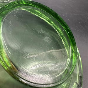 May include: Close-up of a green glass dish with an oval shape. The glass has a slight tint and visible scratches on the surface. The rim of the dish is thicker and more vibrant in color.