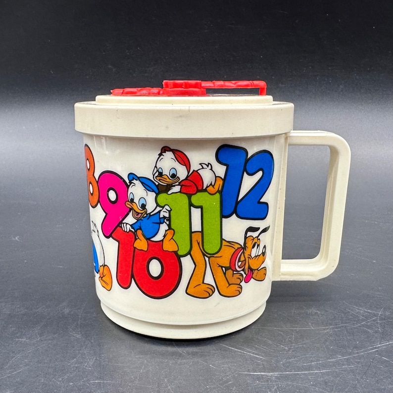 Puede incluir: Una taza de pl&aacute;stico crema vintage con personajes de Disney y los n&uacute;meros 8, 9, 10, 11 y 12 en varios colores. La taza tiene un asa y una tapa roja con un asa. La taza presenta a Donald Duck, Huey, Dewey y Pluto.