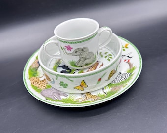 May include: A white ceramic children's dish set featuring a plate, bowl, and handled cup. The set is decorated with a green border and illustrations of various animals, including an elephant, zebra, and leopard.