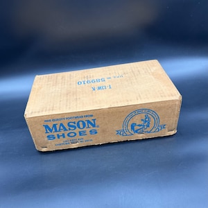 May include: A rectangular cardboard box with the words "MASON SHOES" in blue. The box also has the text "FINE QUALITY FOOTWEAR FROM" and "1251 FIRST AVE CHIPPEWA WI 54729". The box is on a dark blue surface.