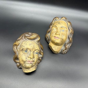 Puede incluir: Dos caras de querubín de yeso antiguo con detalles pintados. Los querubines tienen el pelo castaño, los ojos azules y las mejillas sonrosadas.