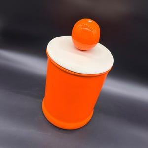May include: A cylindrical orange canister with a smooth matte finish. The container features a flat, circular white lid topped with a glossy, spherical orange handle that resembles a small fruit, providing a contrast in both color and texture.