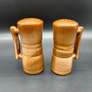 以下が含まれることがあります： 両側にハンドルが付いた、茶色のセラミック製の塩と胡椒入れのセット。シェーカーはテクスチャ加工された釉薬とわずかに丸みを帯びた形をしています。